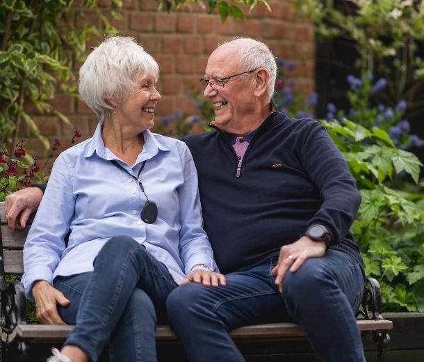 Smiling elderly couple sitting on a bench, the woman wearing a HelpAlert pendant alarm and the man wearing a GPS personal alarm watch for seniors.