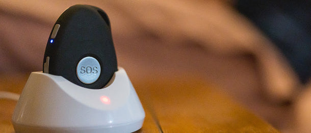 Personal emergency response device with an SOS button resting in a white charging dock on a bedside table, with a blurred figure lying in bed in the background.