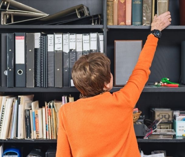 Elderly woman in orange jumper reaching for a book on a shelf while wearing a HelpAlert personal alarm watch