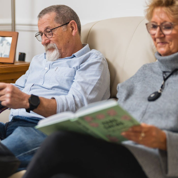 Elderly couple sitting comfortably at home wearing their HelpAlert alarm devices, showing peace of mind and ongoing safety through the annual plan.