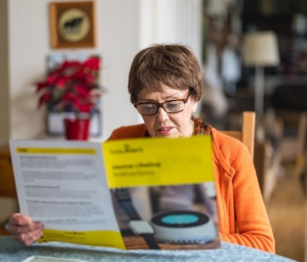 Elderly woman sitting at a table reading a HelpAlert personal alarm brochure at home