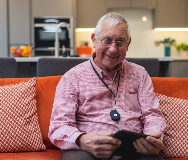 Smiling elderly man sitting on sofa wearing a HelpAlert pendant alarm for safety at home.