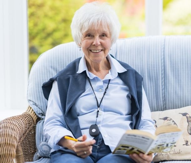 Elderly woman smiling while reading at home, wearing a HelpAlert personal alarm pendant.