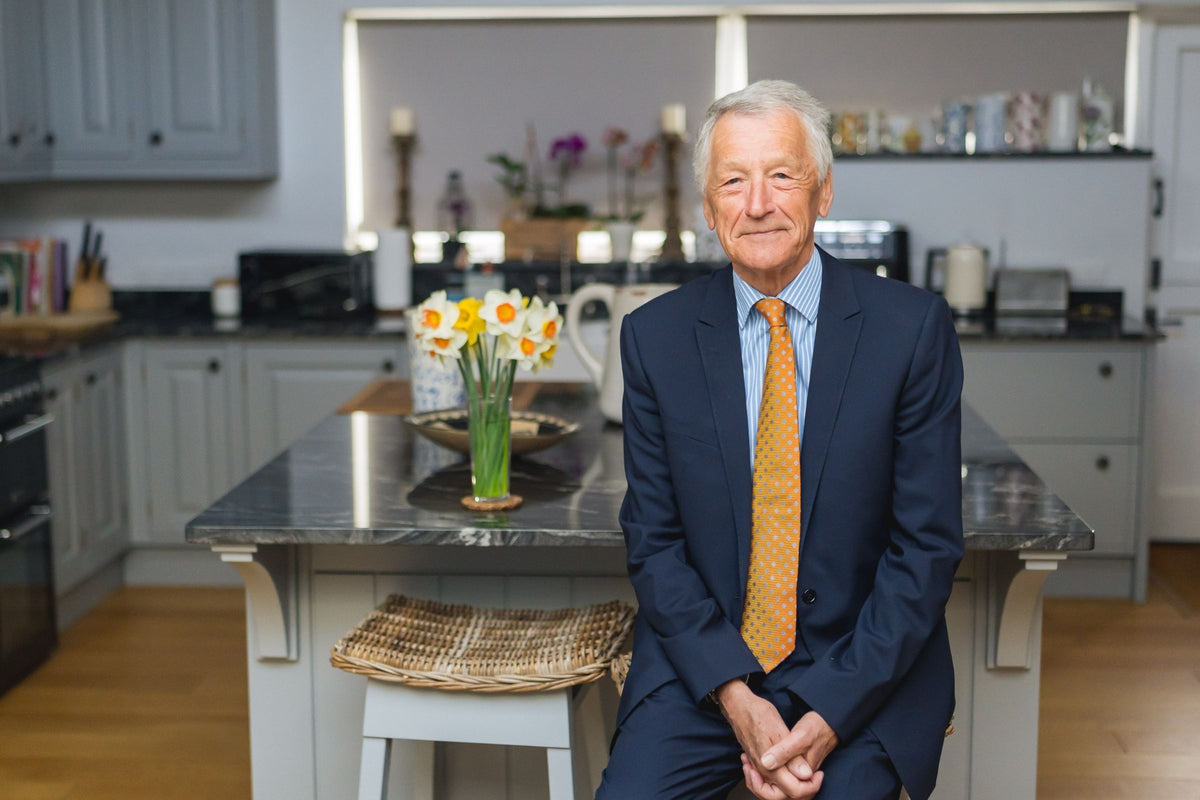 Stewart White seated in modern kitchen wearing navy suit and orange tie